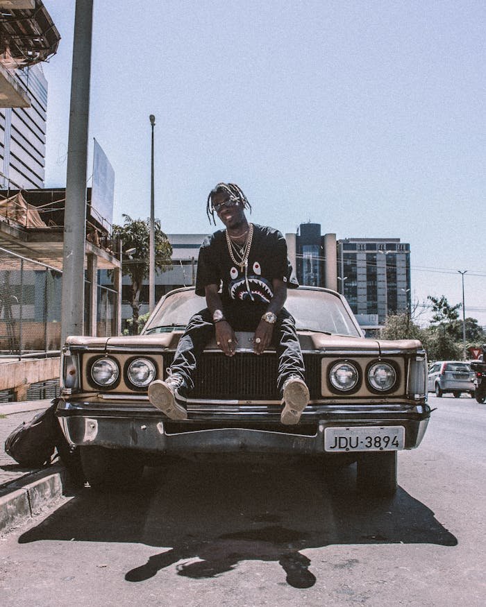 who-we-are Man sitting on vintage car in cityscape, exudes streetwear style and confidence.